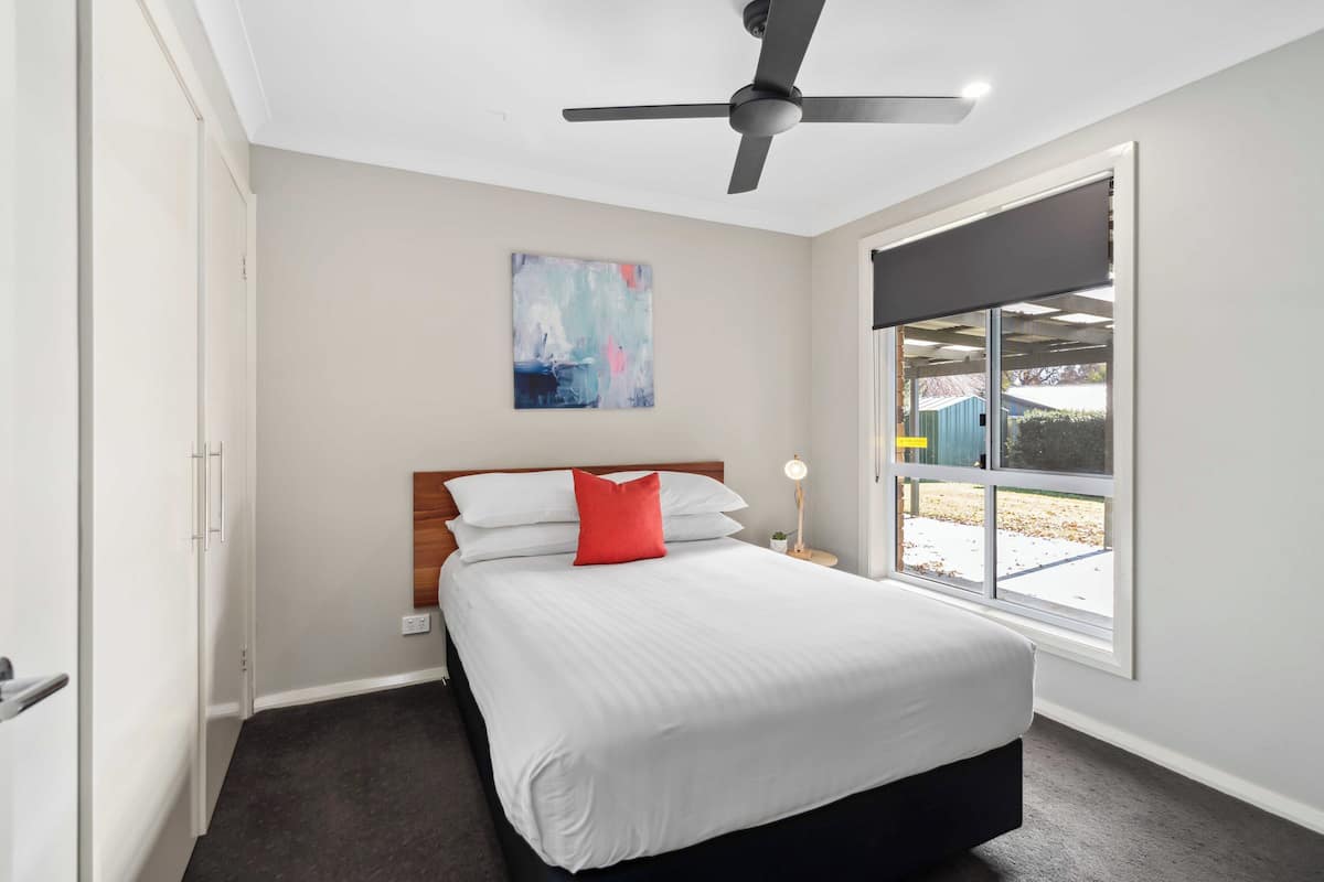 A bedroom with a bed, a ceiling fan, a window with a view, and a painting on the wall.