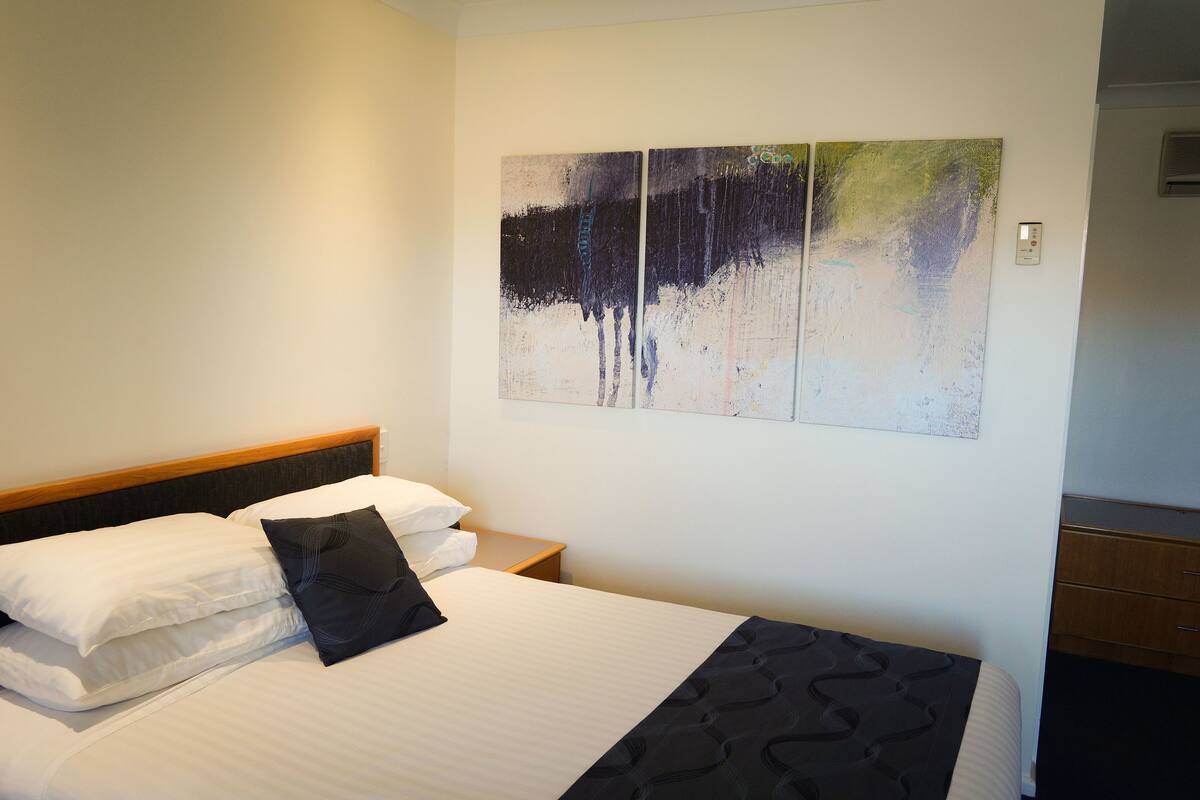 A hotel room with a bed, white linens, a black pillow, a nightstand, and a wall-mounted painting.