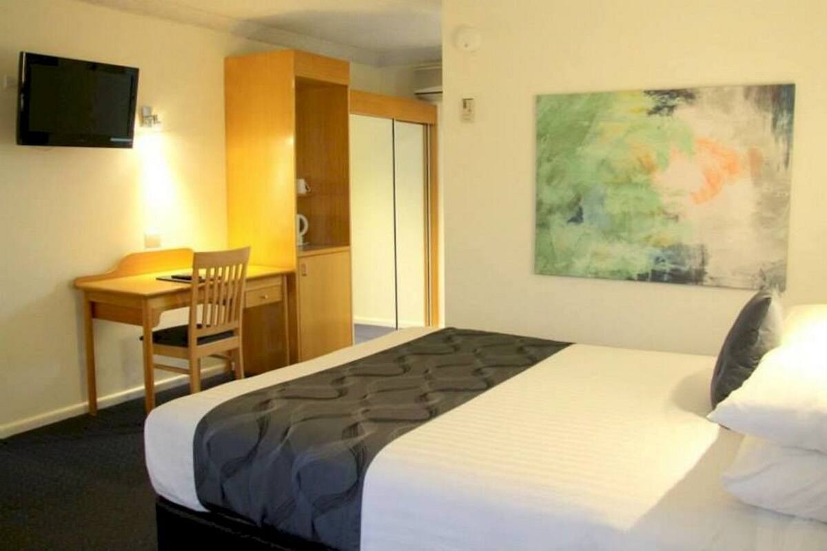 A hotel room with a bed, a desk with a chair, a television, and a painting on the wall.