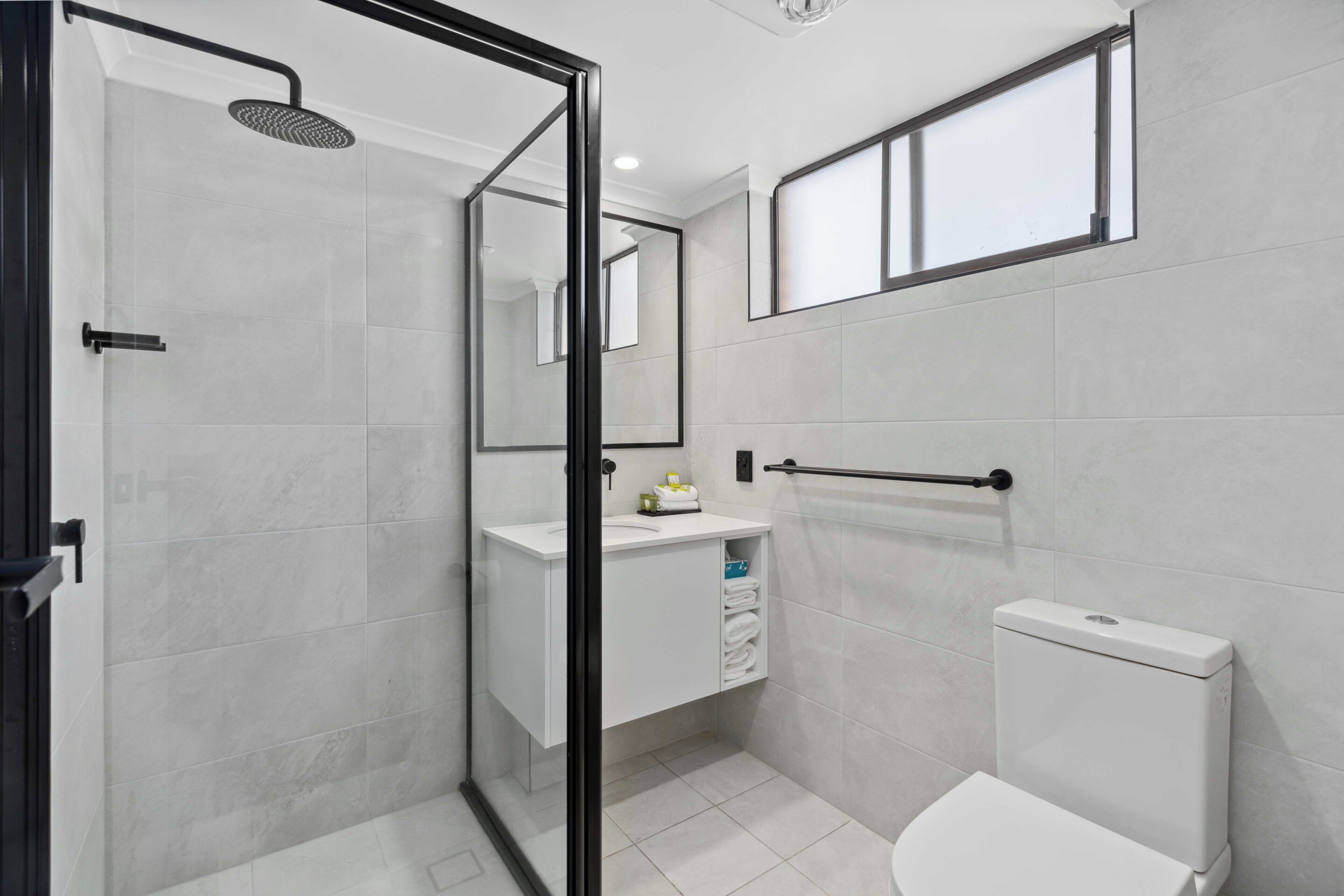 Et moderne badeværelse med et glasafskærmet brusebad, et hvidt toilet og en vask med spejl.