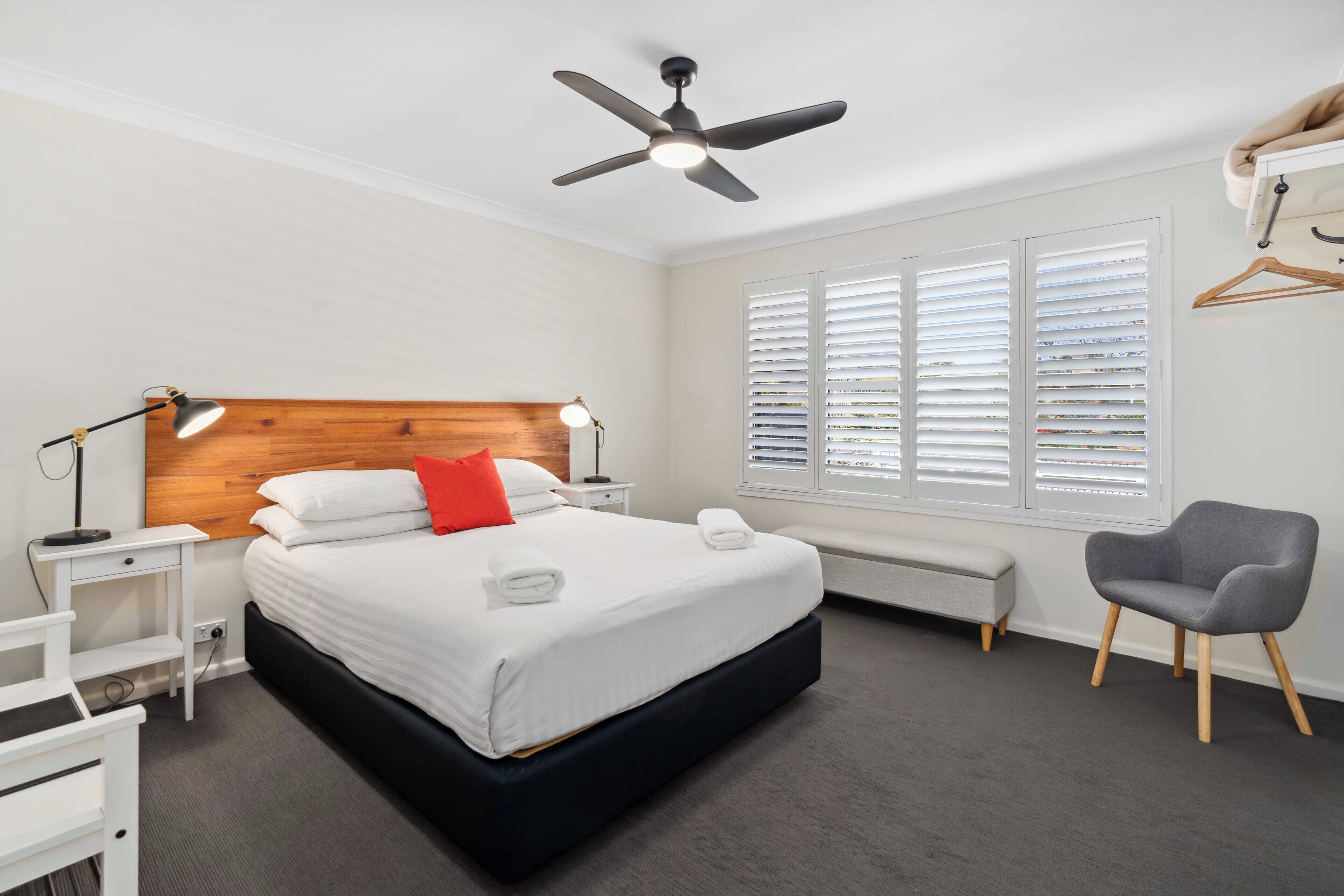 A modern bedroom with a large bed, a ceiling fan, bedside lamps, a bench, and a chair.