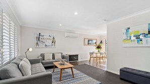 A modern living room with a grey sofa set, a wooden coffee table, and a blue patterned rug. There is a dining area with a table and chairs, and artwork on the walls. - West End Motor Lodge (Orange)