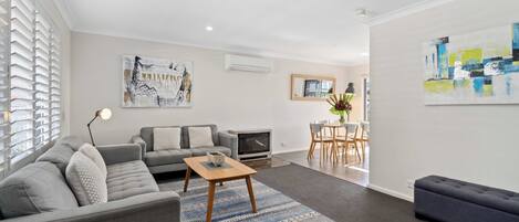 A modern living room with a grey sofa set, a wooden coffee table, and a blue patterned rug. There is a dining area with a table and chairs, and artwork on the walls.