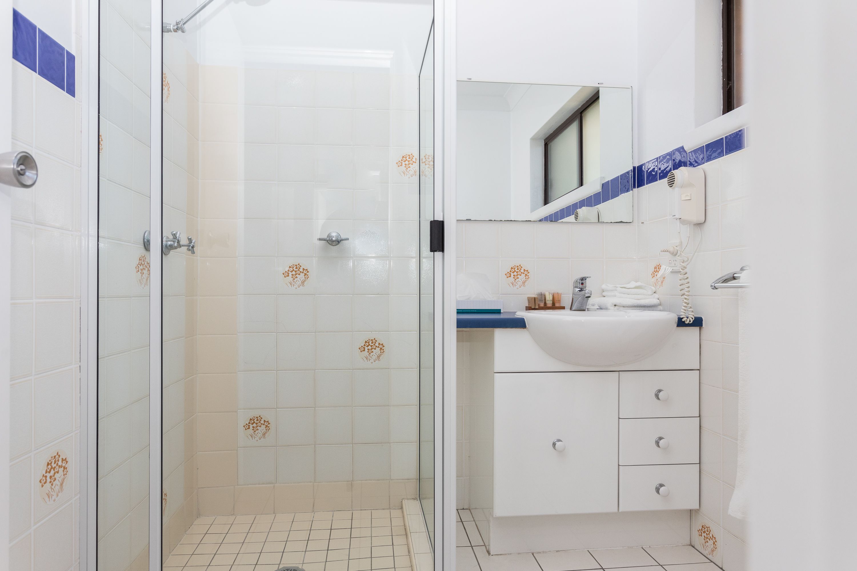 Un baño con cabina de ducha de vidrio, azulejos blancos con motivos florales, un lavamanos blanco y una encimera azul, y un espejo sobre el lavamanos.