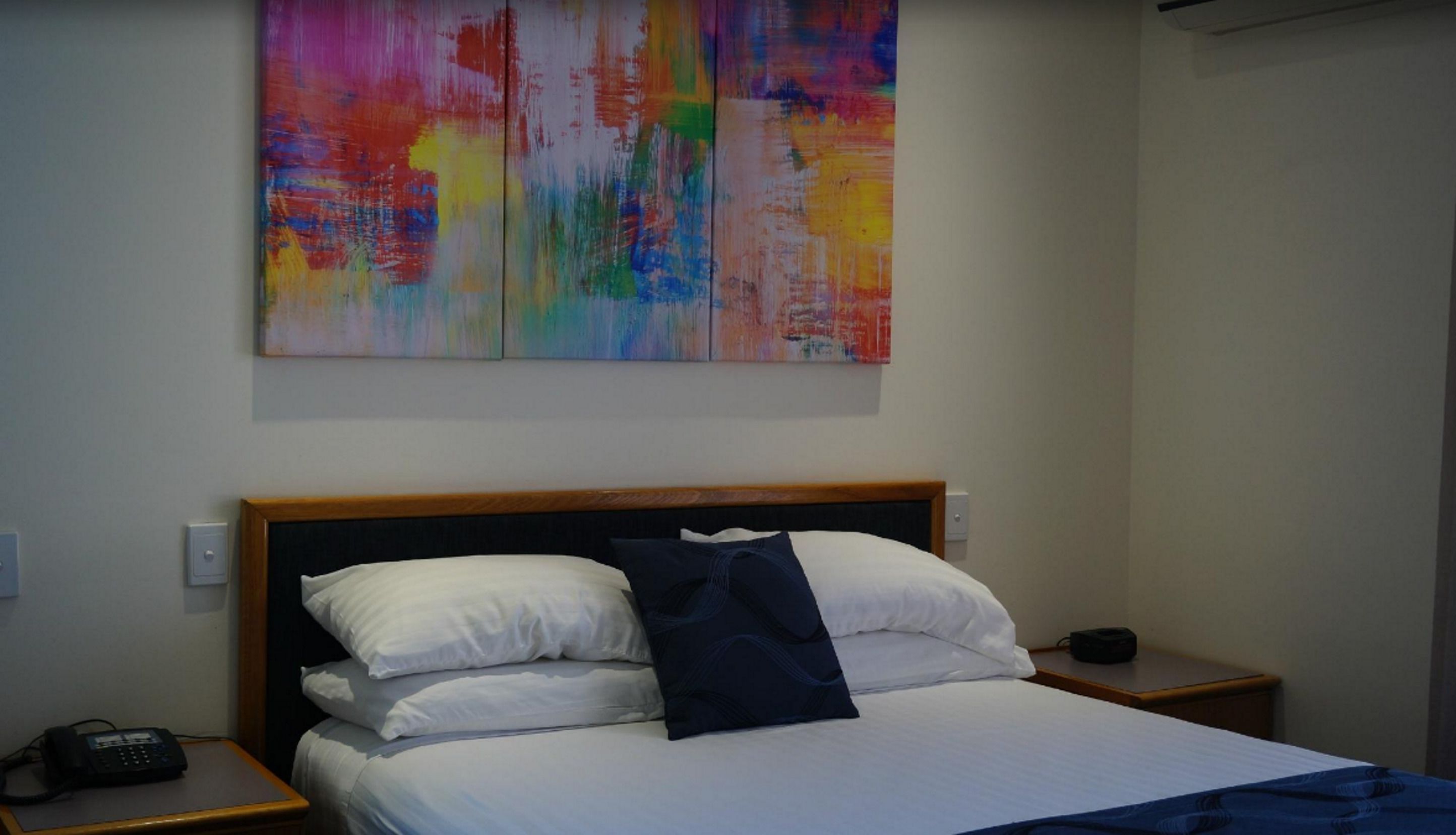 A hotel room with a bed, white bedding, a dark blue pillow, and a colorful abstract painting above the bed. There is a bedside table with a phone and a lamp.