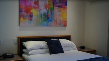 A hotel room with a bed, white bedding, a dark blue pillow, and a colorful abstract painting above the bed. There is a bedside table with a phone and a lamp.