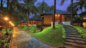 A pathway leading to a building with a thatched roof, surrounded by lush greenery and palm trees. - Coco Lagoon by Great Mount (Anaimalai)