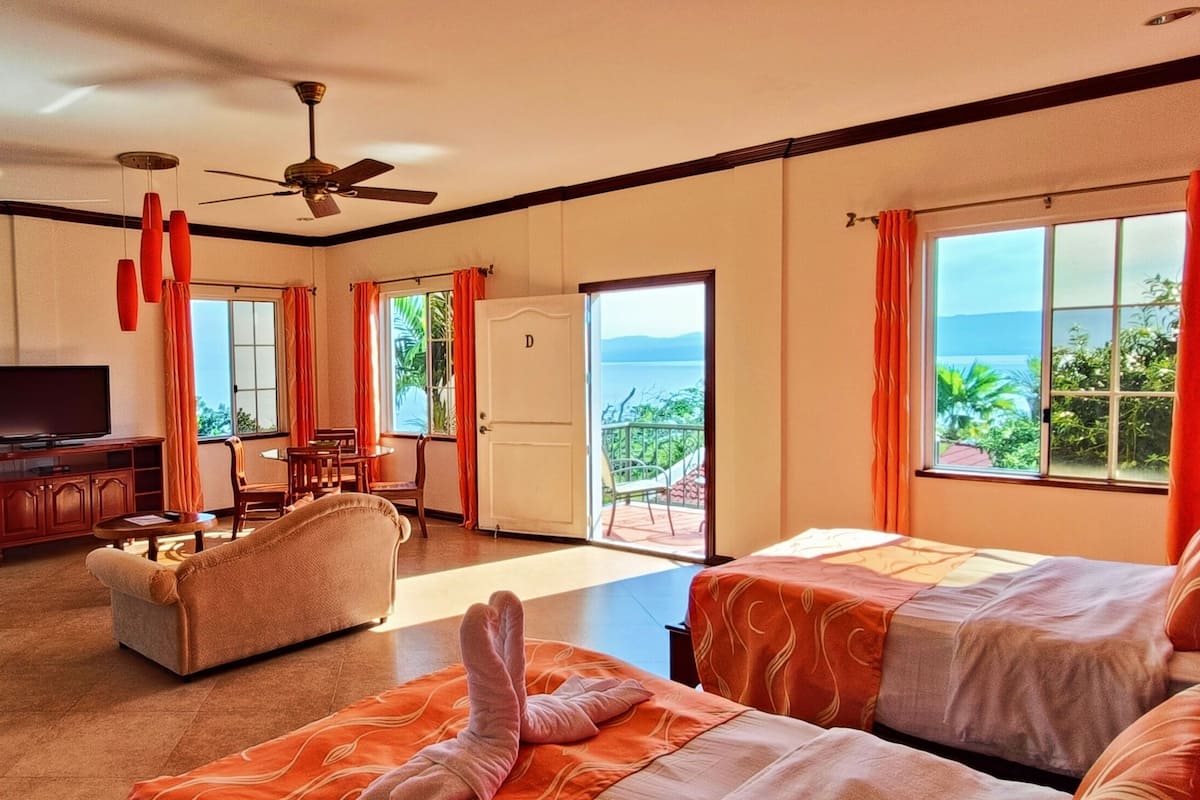 Una habitación de hotel con una cama grande, un sofá, un televisor y un balcón con vista al mar.