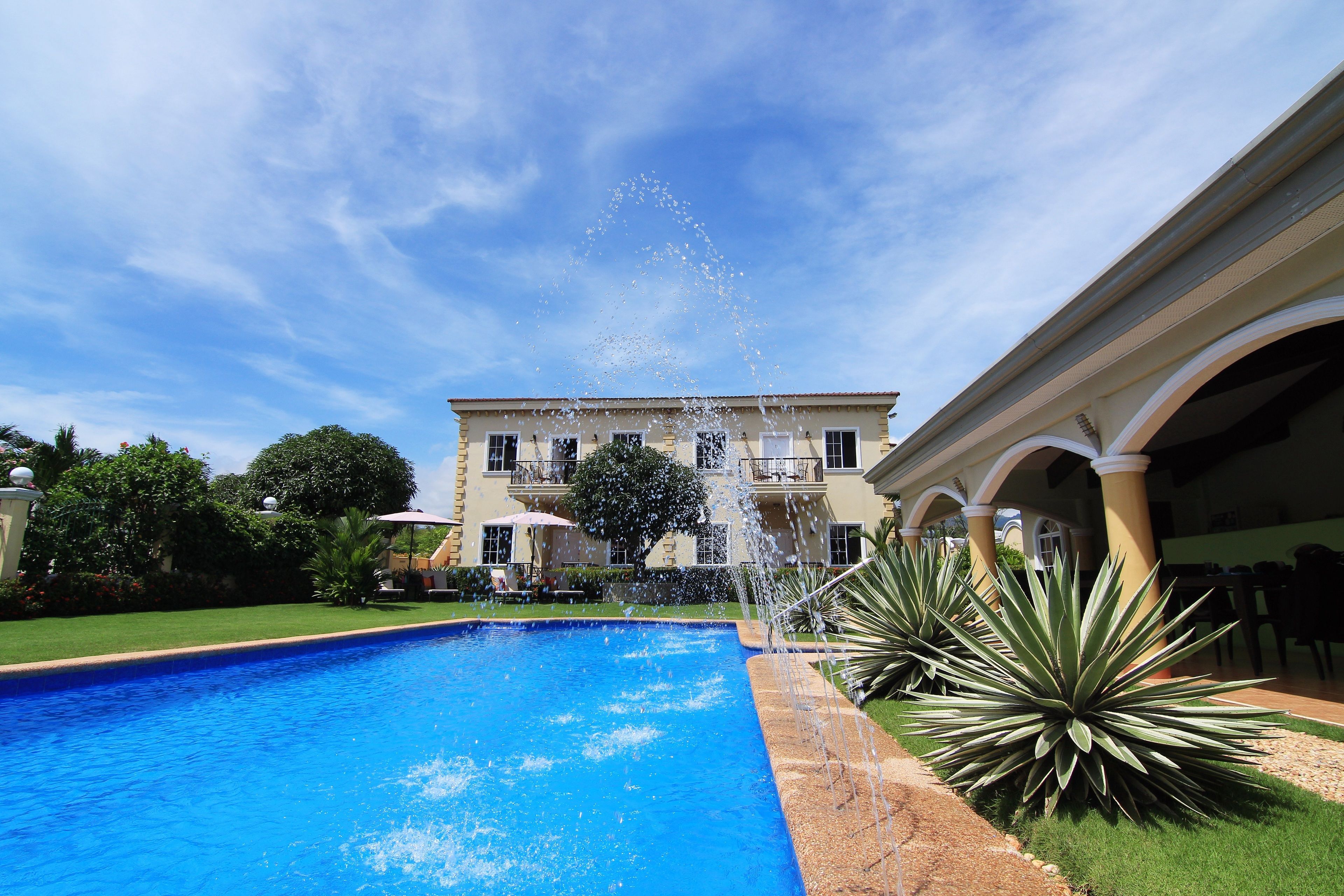 A swimming pool with a water fountain, surrounded by a well-maintained lawn and a large building with multiple balconies.