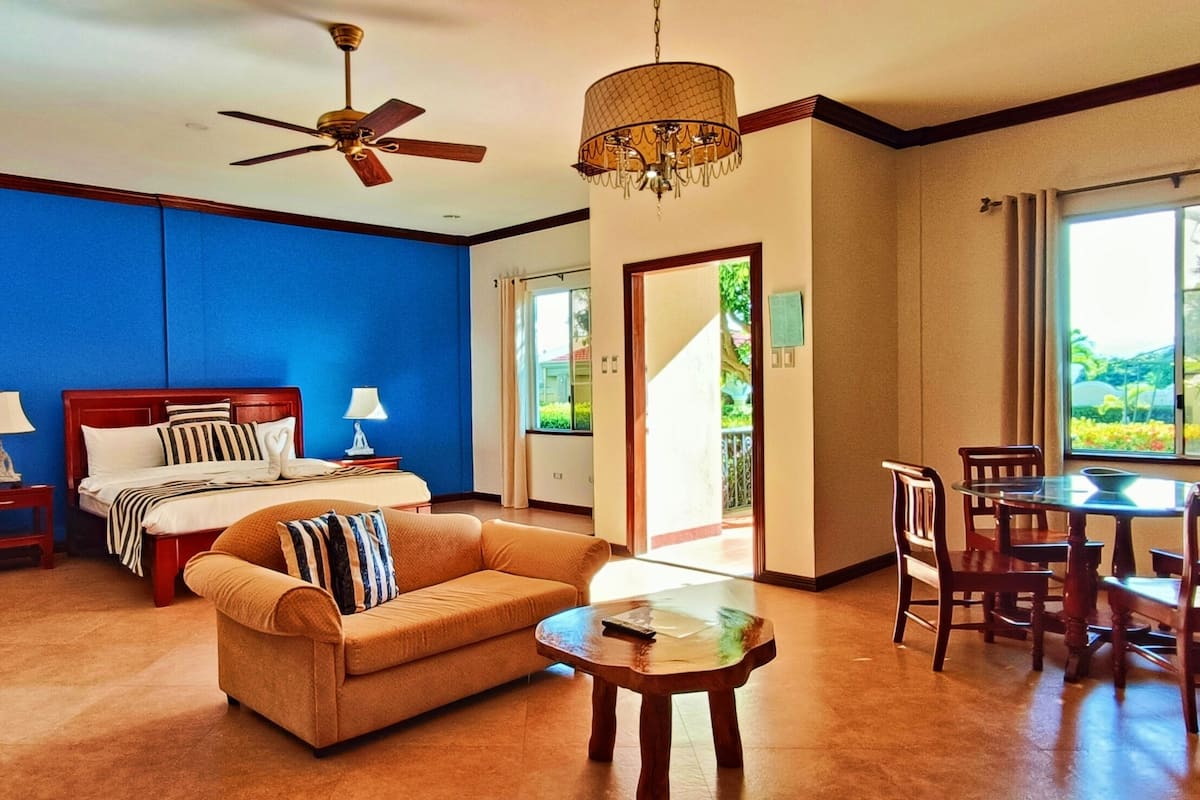 Habitación de hotel con cama, sofá, mesa de comedor con sillas, ventilador de techo y candelabro.