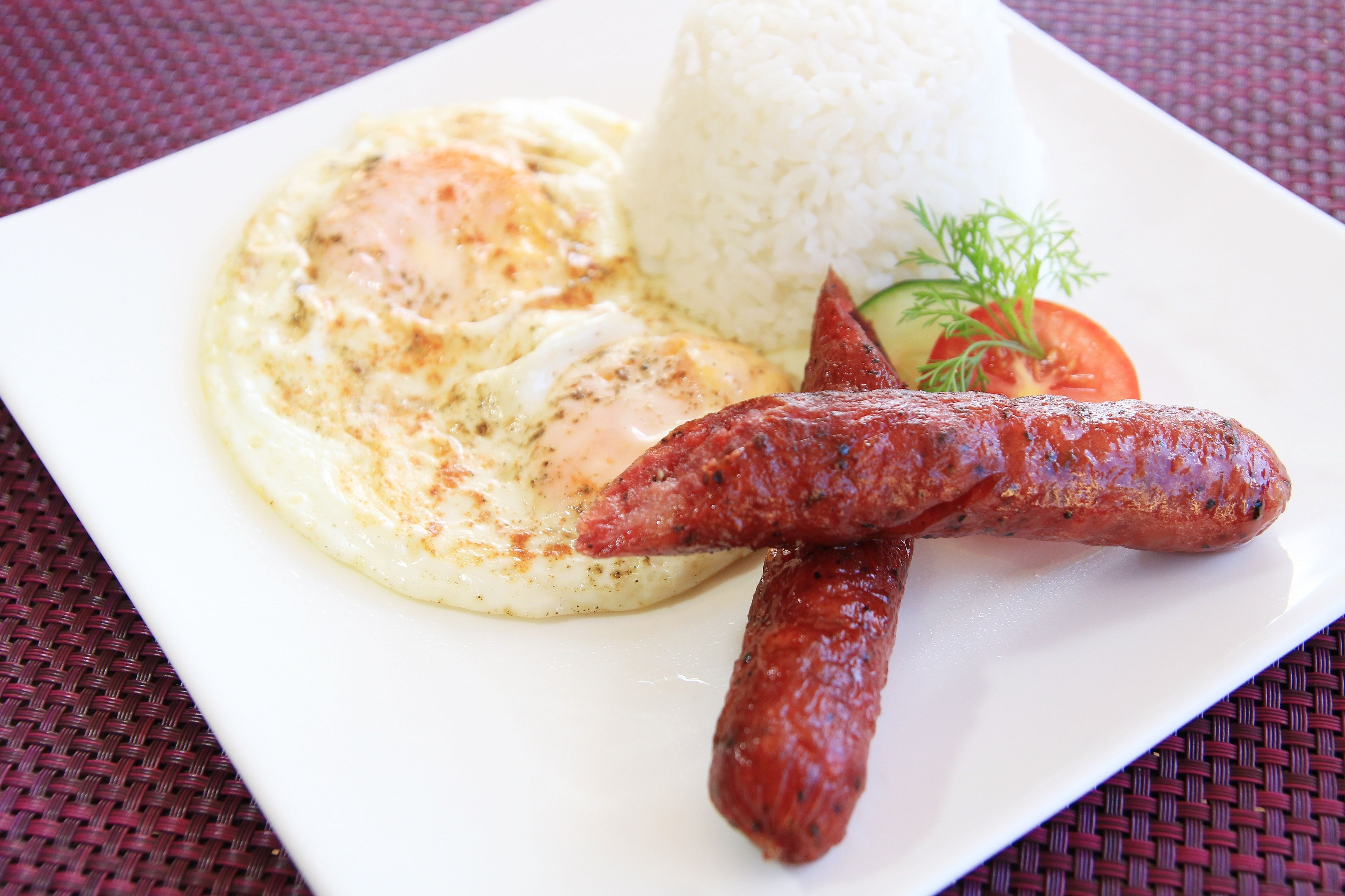 Un plato con huevos revueltos, arroz blanco y salchichas a la plancha, decorado con una rodaja de tomate y una ramita de eneldo.