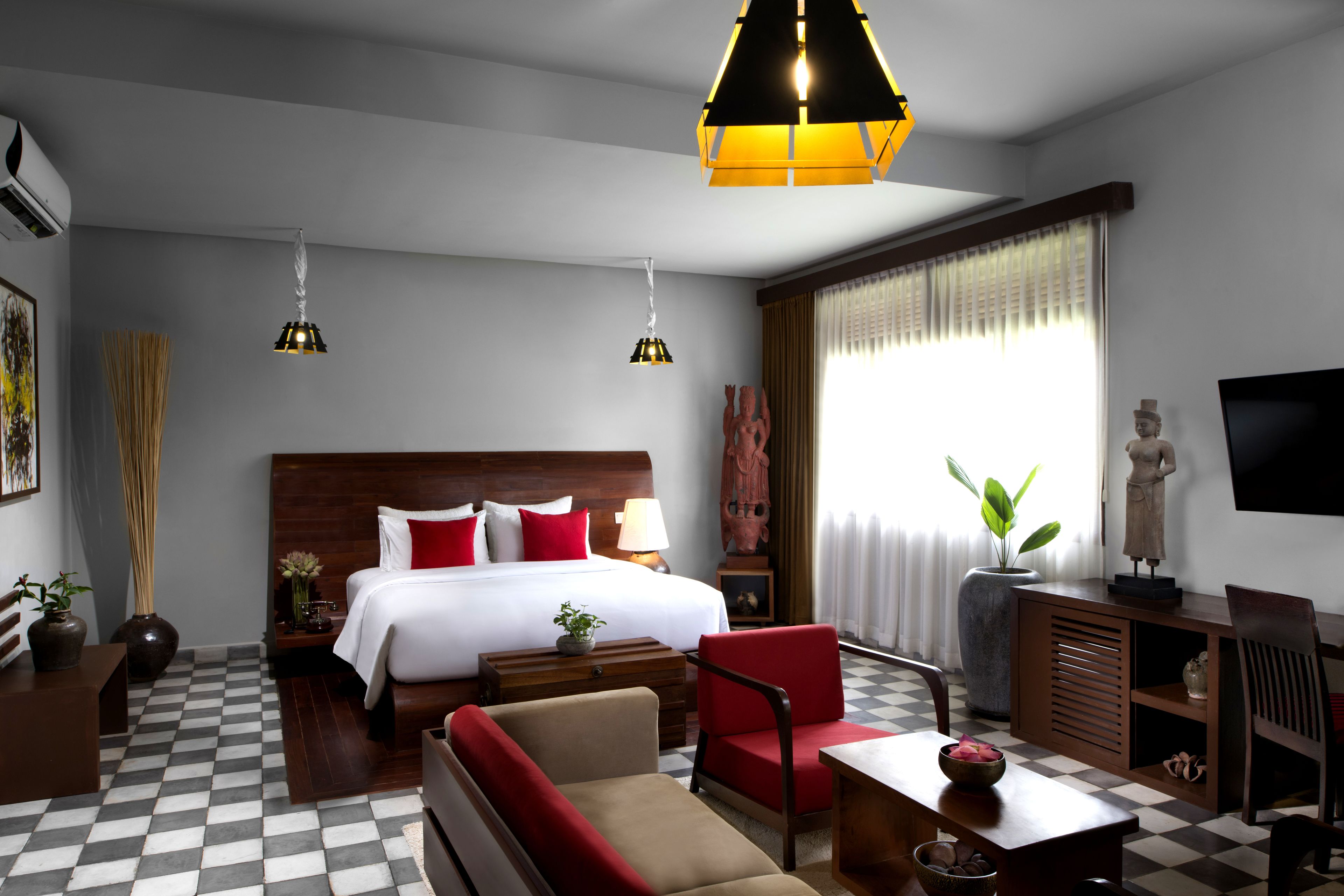 A hotel room with a checkered floor, a bed with white bedding and red pillows, a red armchair, a wooden desk, and a large window with curtains.