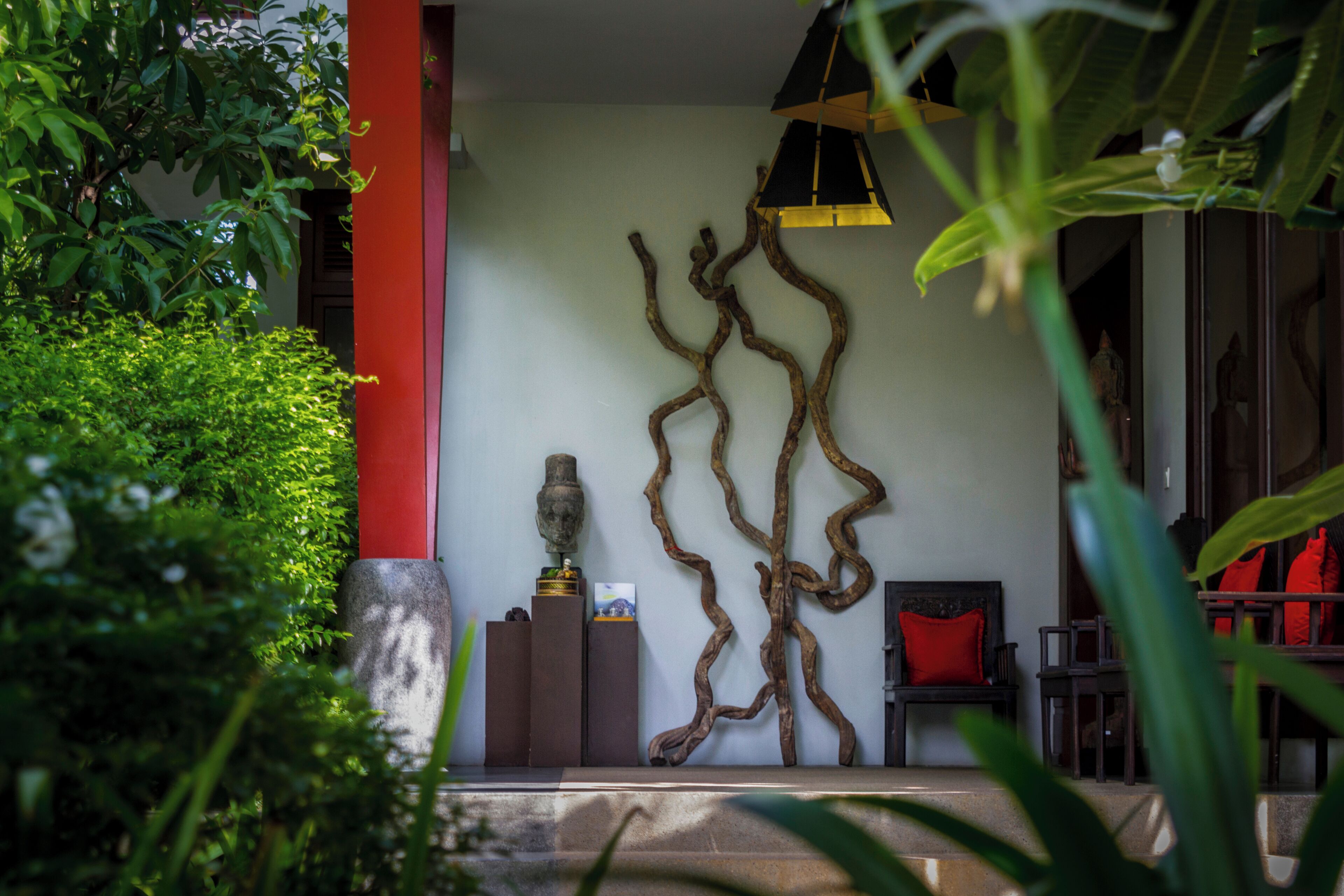 A room with a red pillar, a wall-mounted sculpture, a bench with a red cushion, and a hanging light fixture.