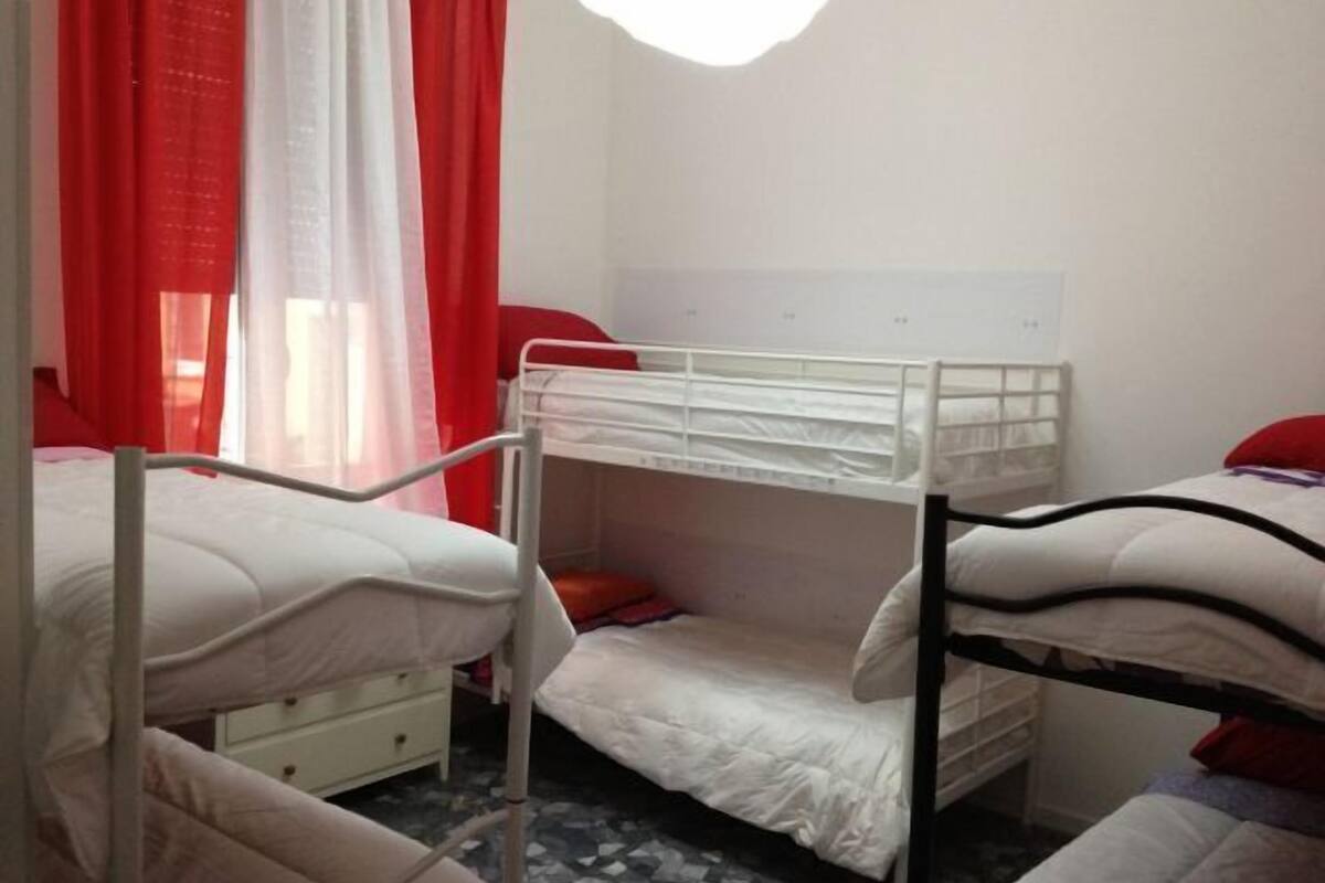 A dormitory room with bunk beds, a white dresser, and a window with red curtains.