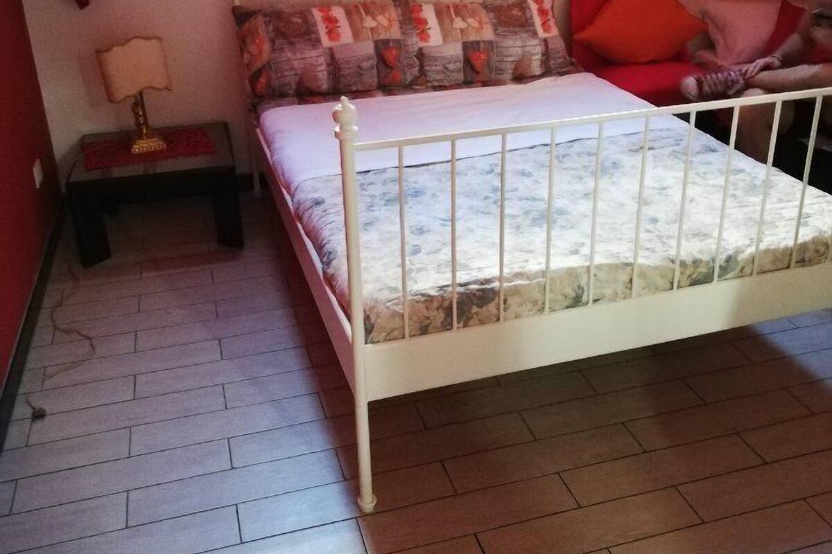 A bedroom with a white metal bed frame, a headboard with a floral design, a nightstand with a lamp, and a red chair.
