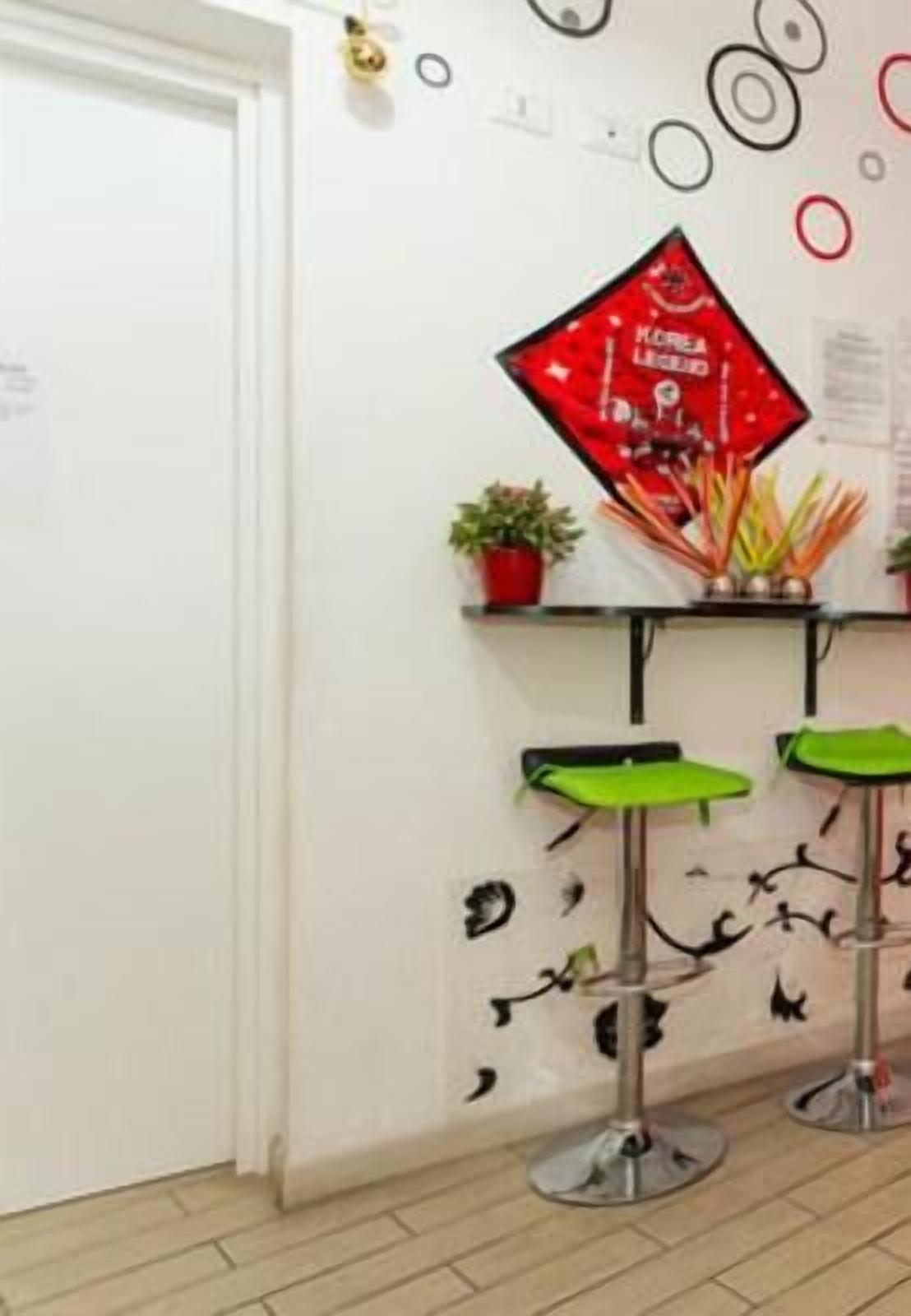 Une pièce avec une porte blanche, un panneau rouge sur le mur, deux tabourets de bar verts et une petite plante en pot.