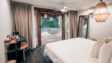 A bedroom with a large bed, a desk, and a view of a garden with a hammock.