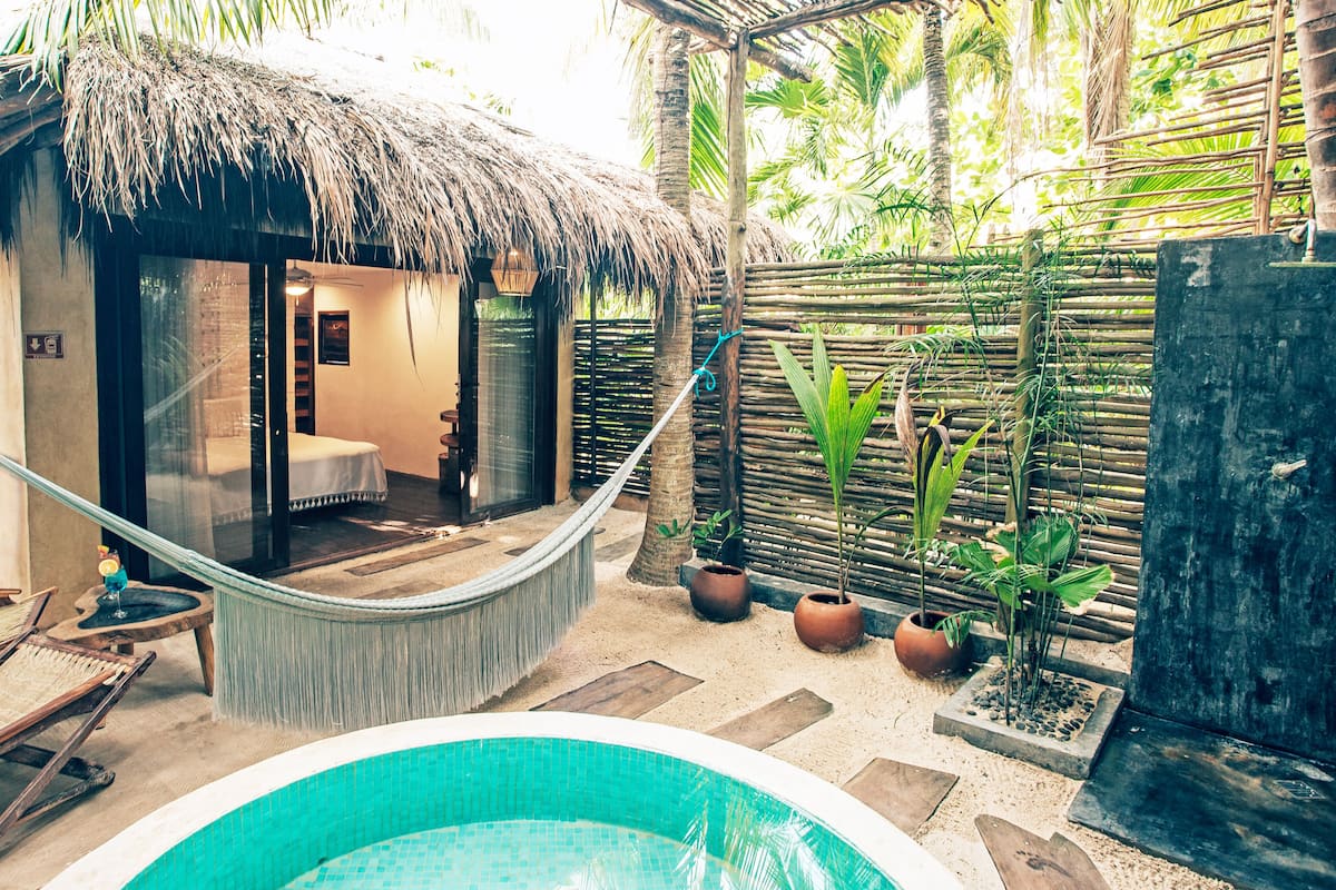 Eine reetgedeckte Hütte mit Glastür zu einem Badezimmer, einer Hängematte, einem Pool und tropischen Pflanzen.