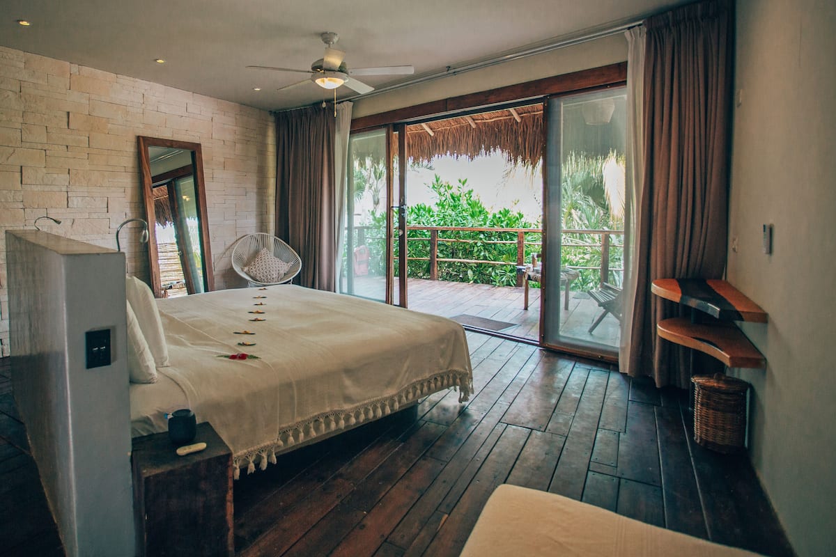 Ein Schlafzimmer mit einem großen Bett, einem Deckenventilator, Holzboden, einem Balkon mit Blick ins Grüne und einer Ziegelwand.