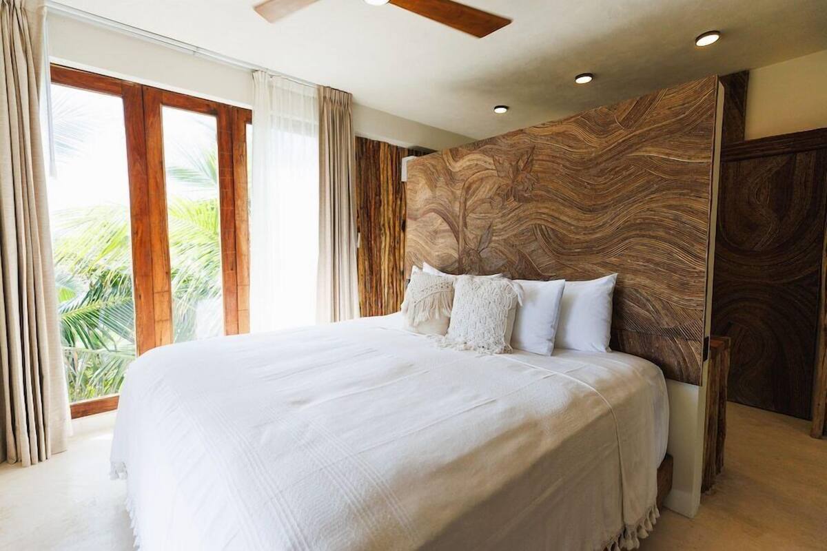 Ein großes Bett mit weißer Bettdecke, ein Holz-Kopfbrett mit Blattmuster und Blick auf Palmen durch das Fenster.