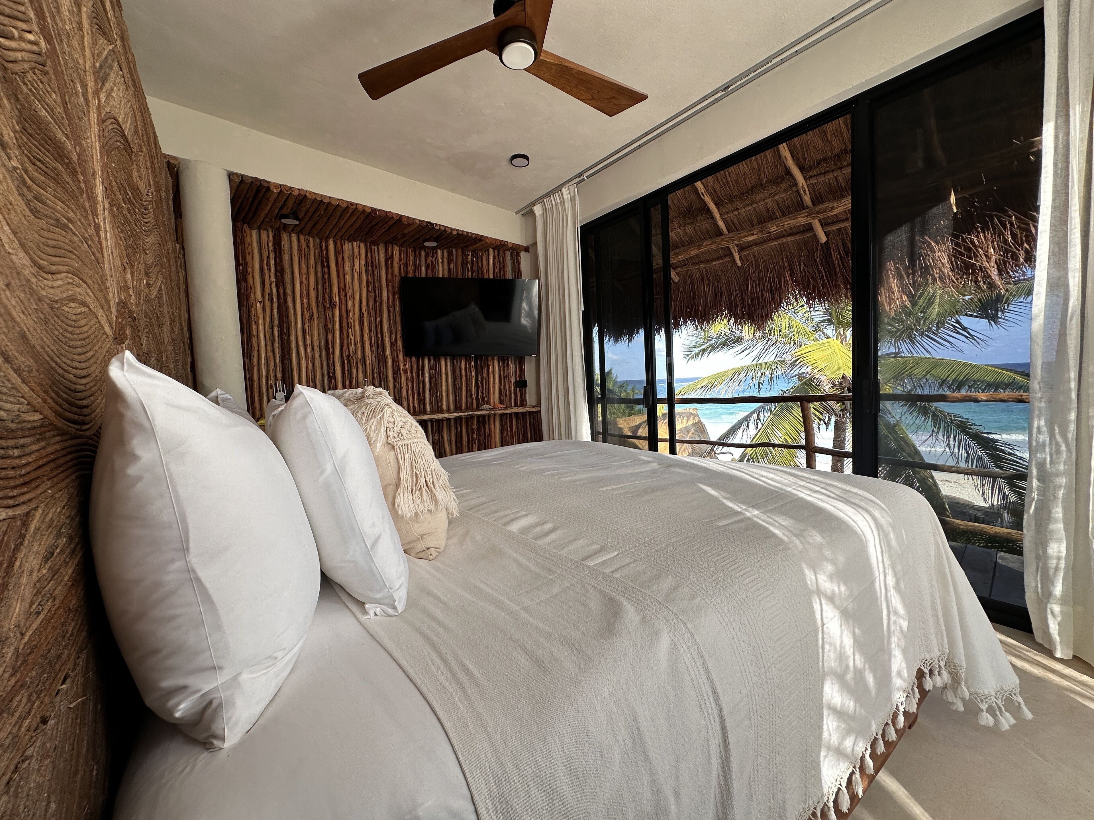 Quarto com uma cama grande, um ventilador de teto, uma TV fixada em uma parede de madeira e vista para o oceano através de amplas janelas.