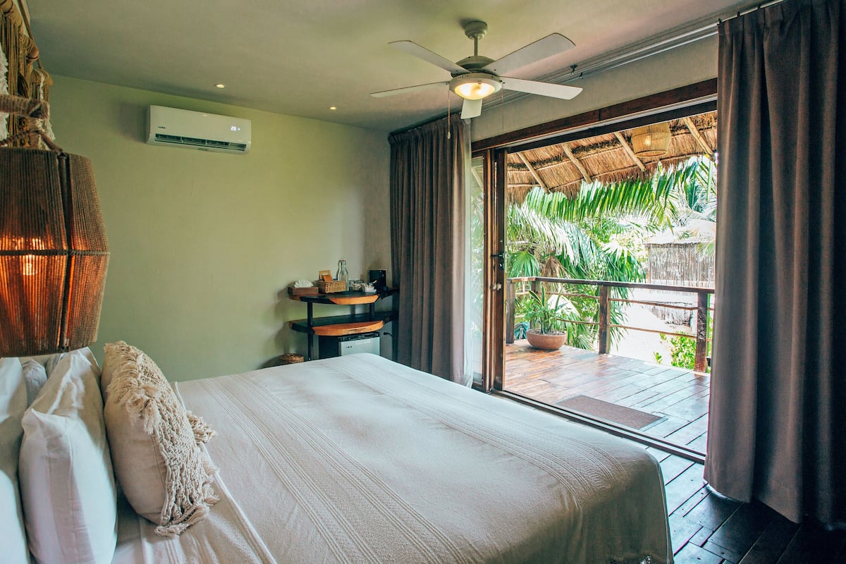 Ein Schlafzimmer mit Bett, Deckenventilator, Klimaanlage, einem kleinen Tisch mit Gegenständen und Blick ins Grüne durch ein Fenster.