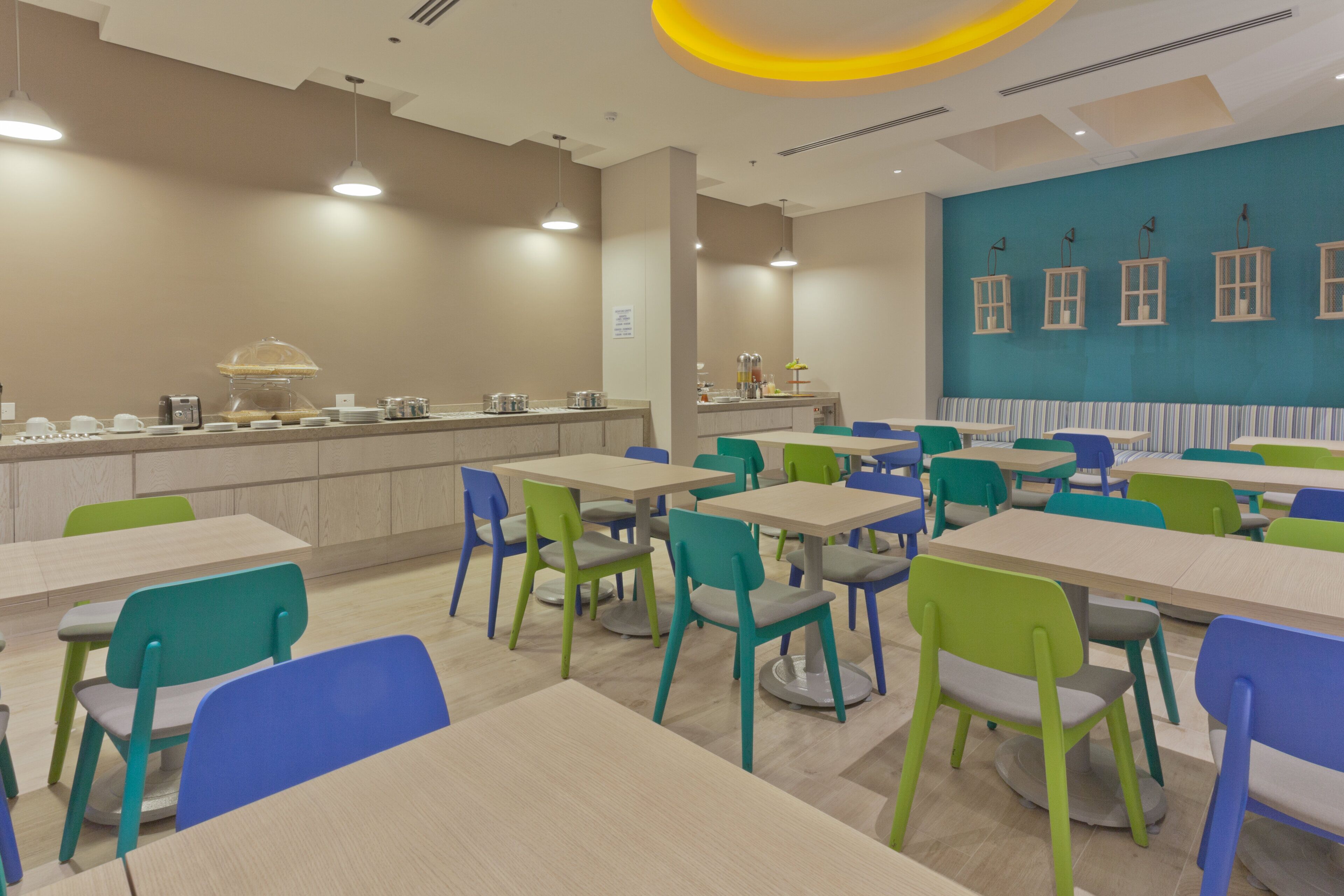 Una cafetería moderna con varias mesas y sillas, una barra con equipo para servicio de alimentos, y una pared azul decorada con motivos de ventanas.