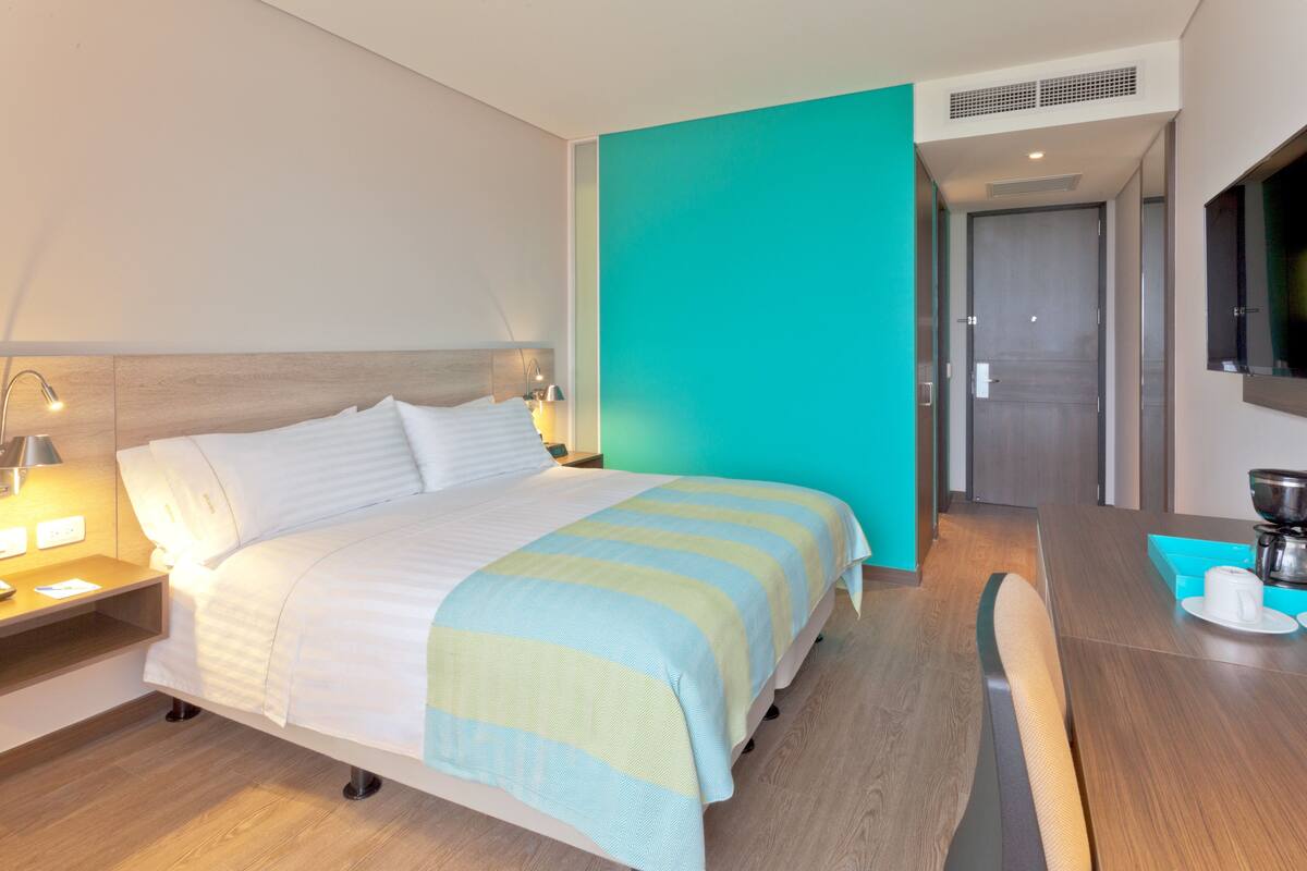 Quarto de hotel com cama, mesa de cabeceira, abajur e máquina de café sobre uma mesa de madeira.