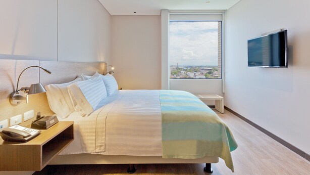 Una habitación de hotel moderna con una cama grande, una mesita de noche con lámpara, un televisor de pantalla plana en la pared y una ventana con vista a la ciudad.