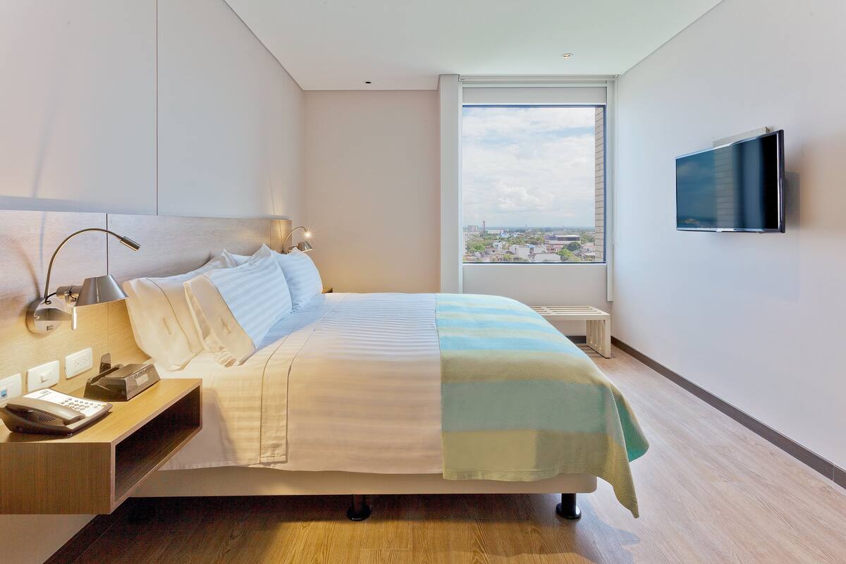 Quarto de hotel moderno com cama grande, mesa de cabeceira com luminária, TV de tela plana na parede e janela com vista para a cidade.