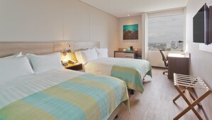 Habitación de hotel con dos camas, un escritorio, una silla, un televisor y ventana con vistas a la ciudad.