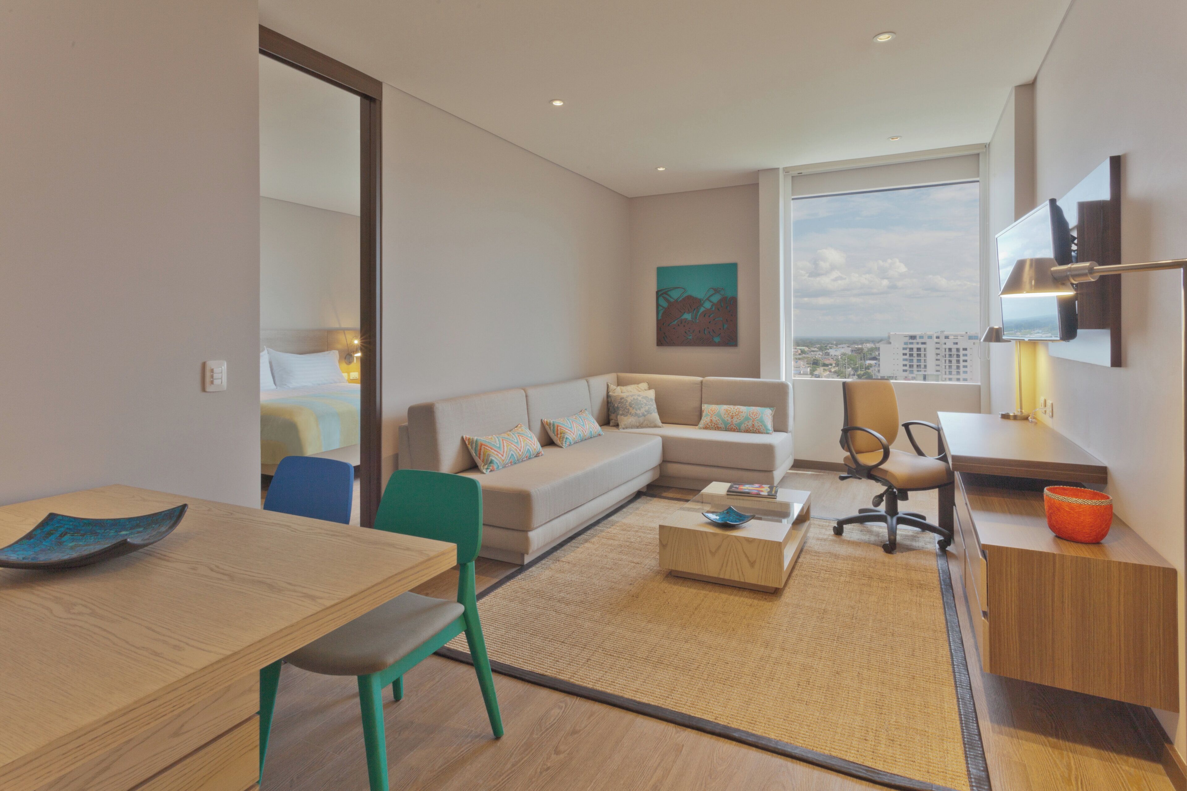 A modern hotel room with a sofa, a dining table, a desk, and a large window offering a city view.