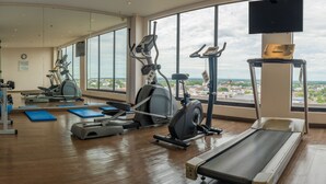 Un gimnasio con diverso equipamiento de ejercicio, amplias ventanas y vistas a un paisaje urbano.