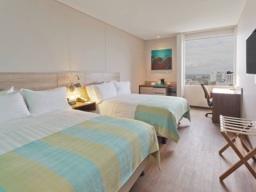 A hotel room with two beds, a desk, a chair, a TV, and a window with a city view.