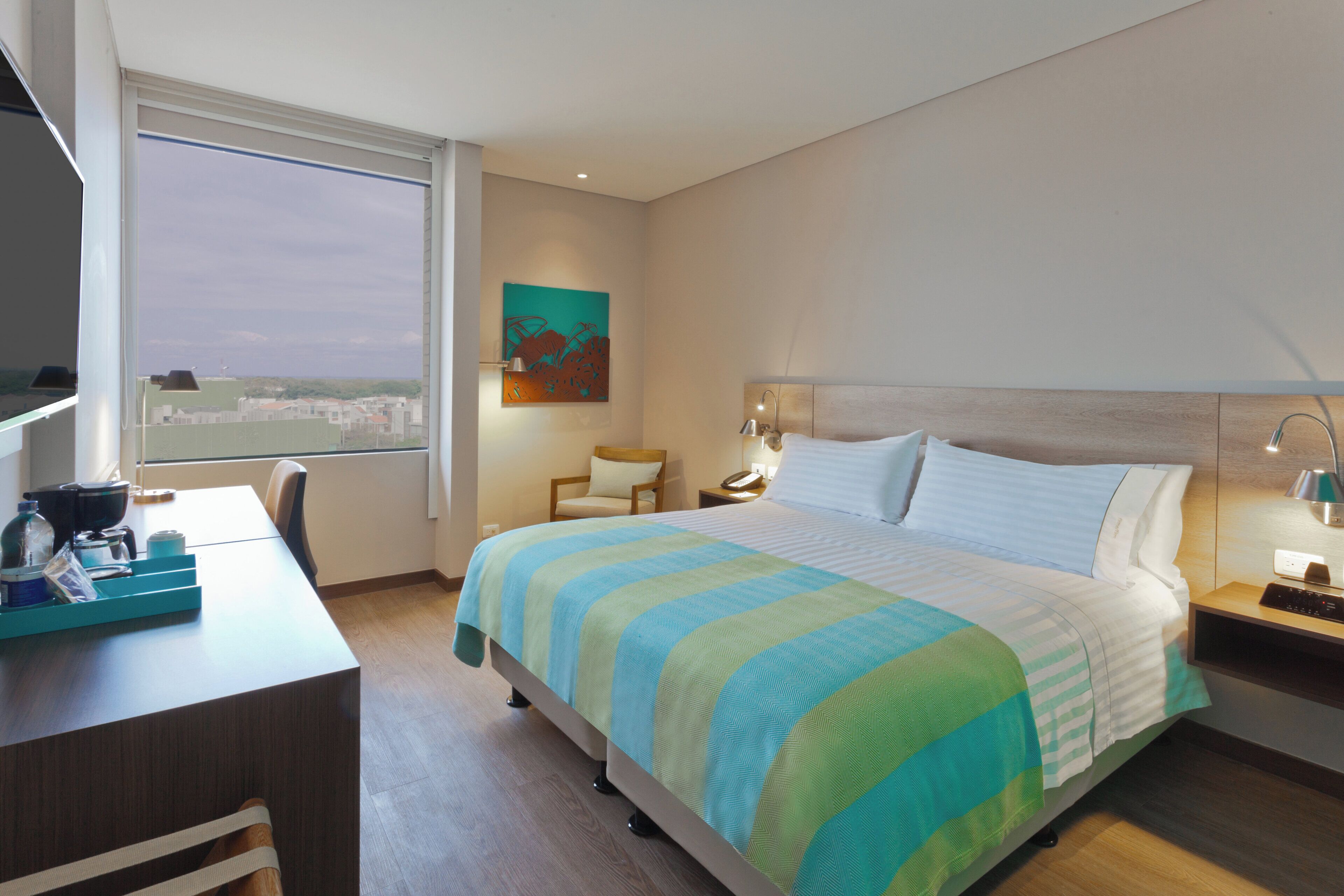 Habitación de hotel con cama, escritorio, silla, ventana y un cuadro en la pared.