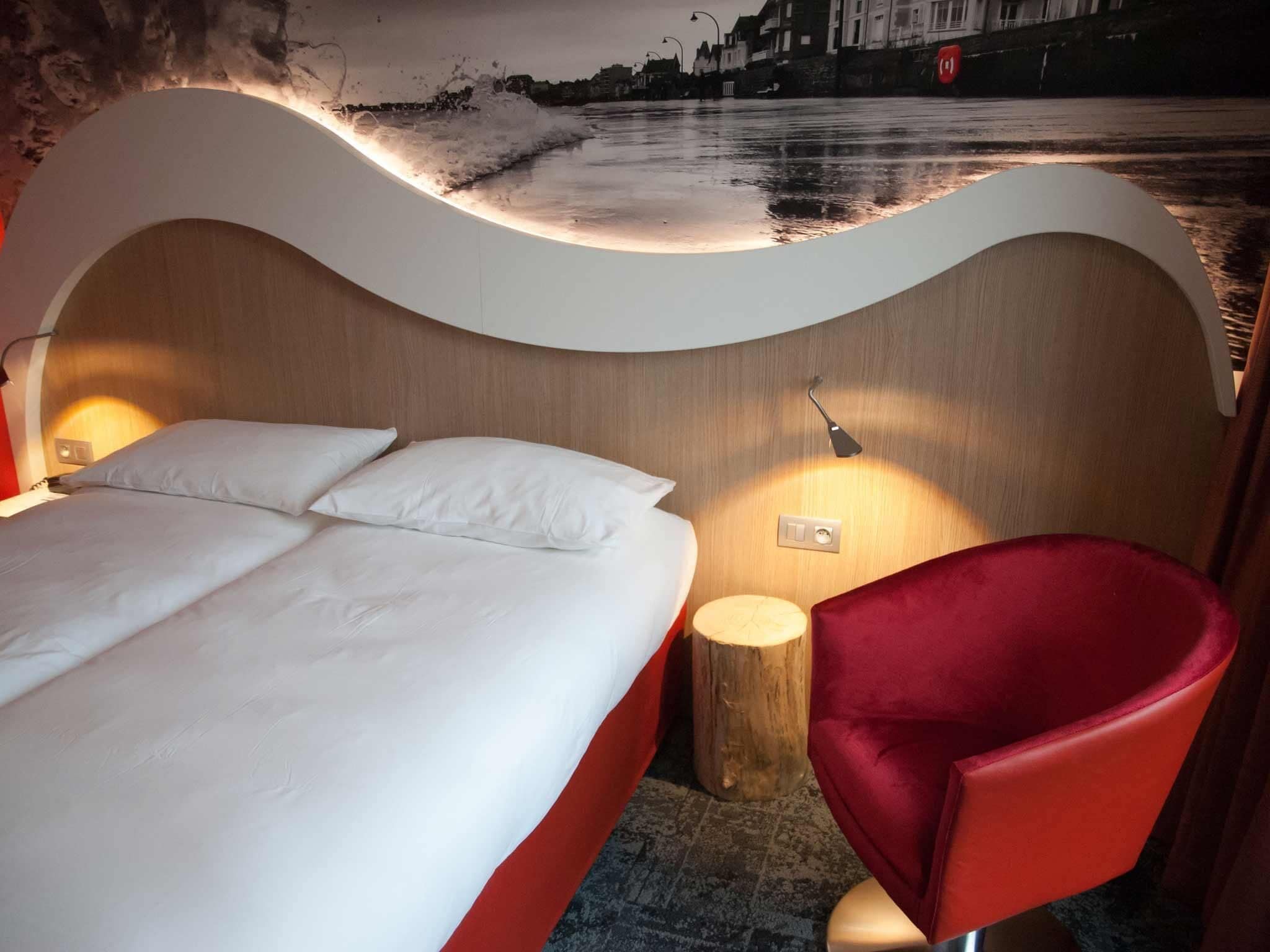 Een moderne hotelkamer met een gebogen hoofdeinde, een bed met witte lakens, een rode stoel en een nachtkastje met een lamp.