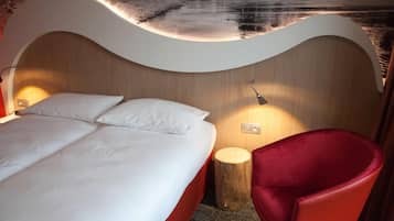 Une chambre d’hôtel moderne avec une tête de lit incurvée, un lit recouvert de draps blancs, une chaise rouge et une table de chevet avec une lampe.