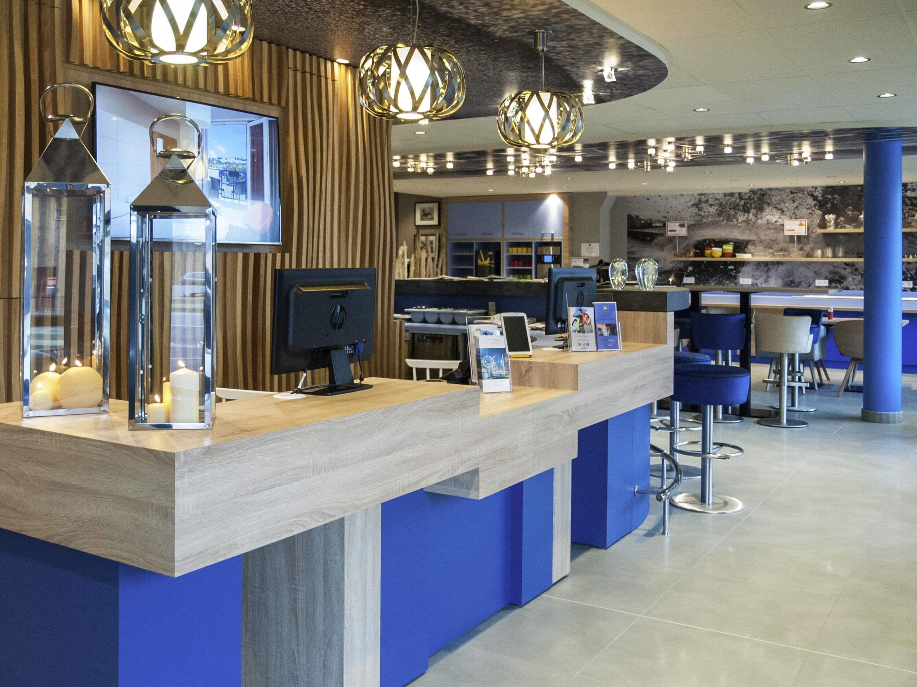 Un comptoir d’accueil moderne, doté d’un ordinateur, d’un mur recouvert de panneaux en bois et d’un espace bar en arrière-plan.