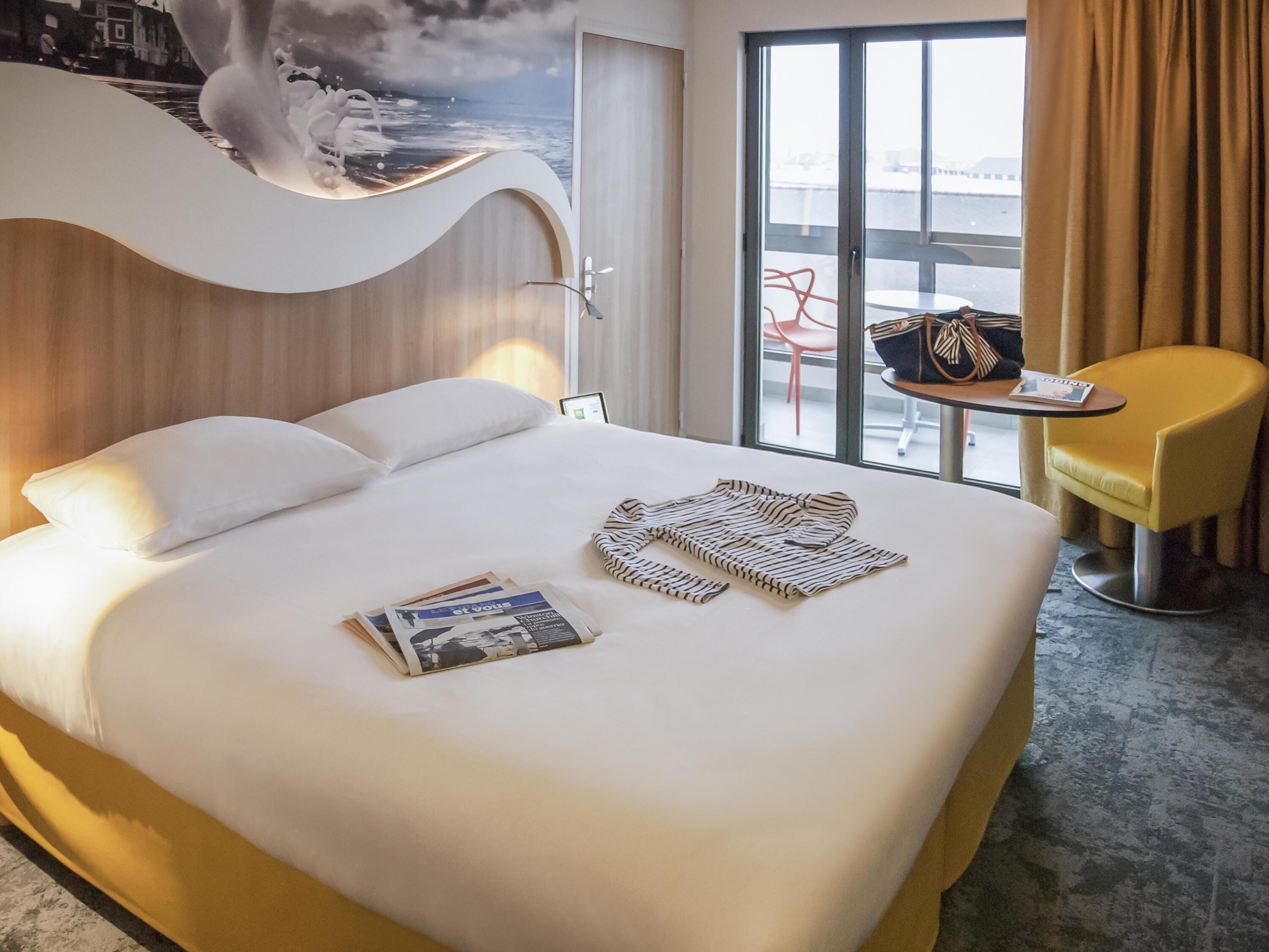 Habitación de hotel con una cama grande, una mesita con una silla, vistas al exterior a través de un ventanal amplio y una pared con un diseño original.