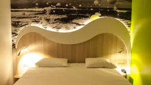 Un dormitorio con cabecera curva, cama con dos almohadas, mesita de noche con lámpara y un mural en la pared con un mar embravecido.