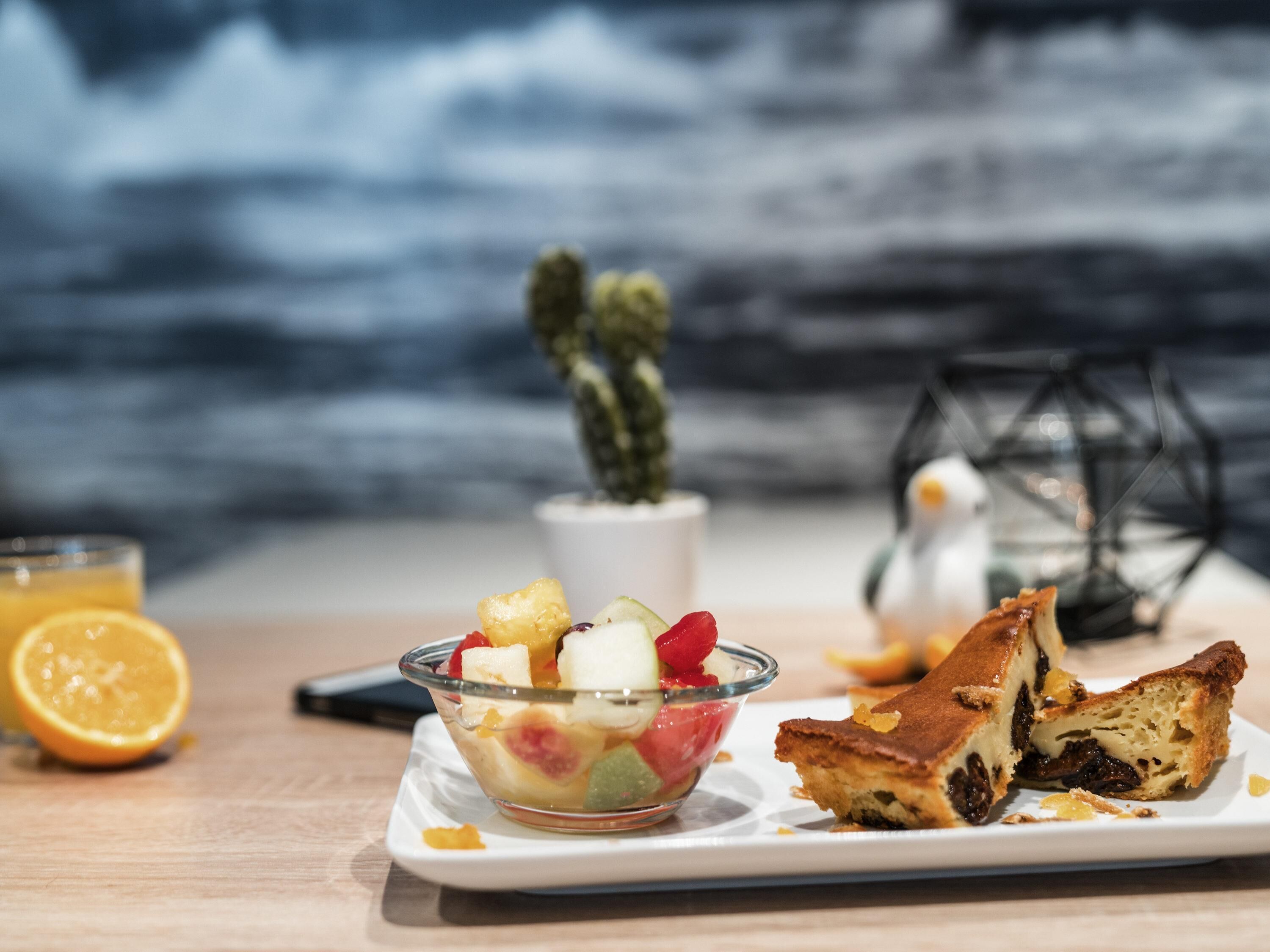 Une assiette de dessert avec des fruits et un morceau de gâteau, un verre de jus d’orange et une plante succulente (un cactus).