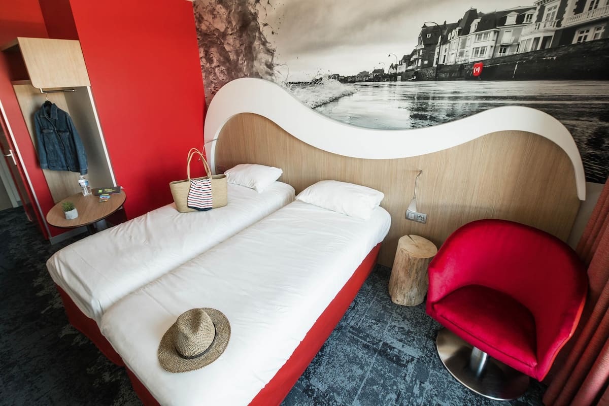 A hotel room with a bed, a red chair, a small table with a lamp, a wall with a cityscape mural, and a red wall.