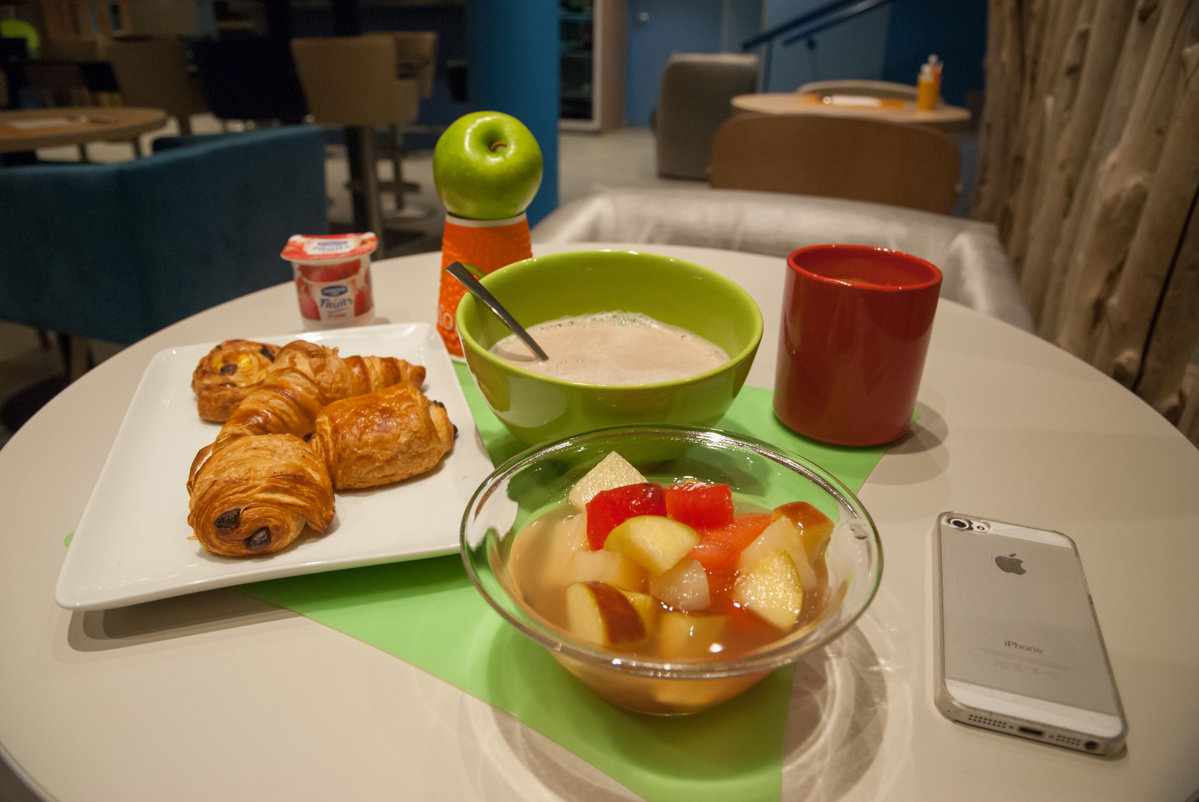 A breakfast setting with croissants, a bowl of fruit, a red cup, a green bowl with a spoon, and a smartphone on a table.