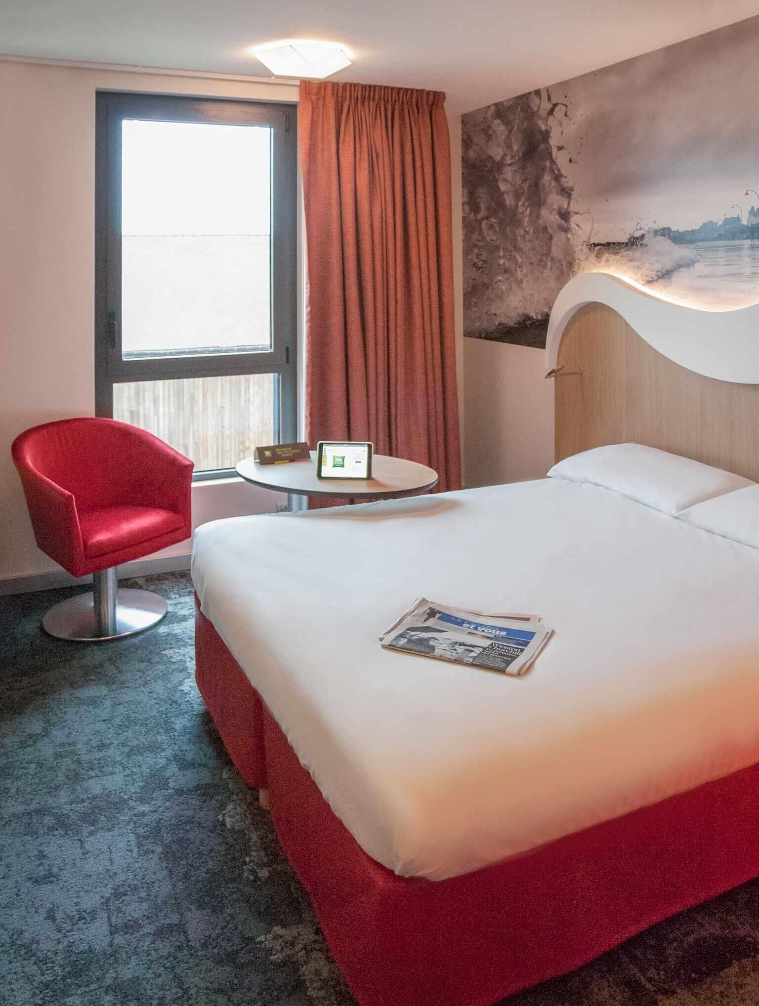 Hotellihuone, jossa on sänky, punainen tuoli, pieni pöytä ja ikkuna, jossa on verhot.