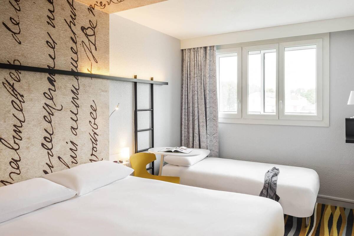 Habitación de hotel con dos camas, una ventana con cortinas, un escritorio con una silla y una pared con detalles decorativos escritos.