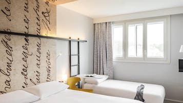 A hotel room with two beds, a window with curtains, a desk with a chair, and a wall with decorative script.