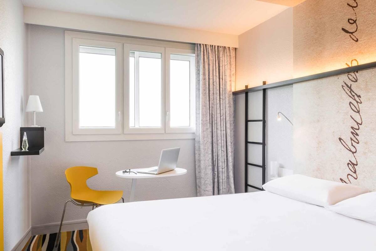 Habitación de hotel con cama, escritorio, computadora portátil, silla, ventana y un elemento decorativo en la pared.