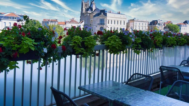 Terraza a orillas del río con vistas a edificios históricos, una valla blanca y mobiliario para exteriores.