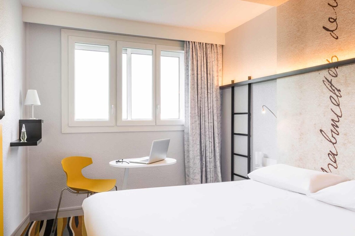 A hotel room with a bed, a desk with a laptop, a chair, a window, and a wall with a decorative element.