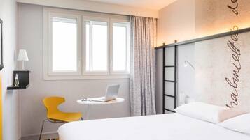 A hotel room with a bed, a desk with a laptop, a chair, a window, and a wall with a decorative element.